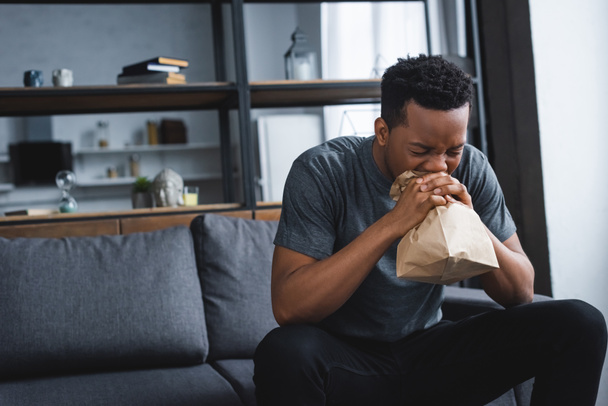 στρεσαρισμένος Αφροαμερικάνος που αναπνέει με χάρτινη σακούλα ενώ έχει κρίση πανικού στο σπίτι - Φωτογραφία, εικόνα