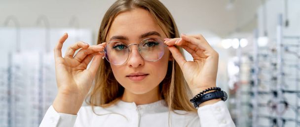 Woman in spectacles. Choosing glasses. Client in optics. Selective focus. Woman in corrective glasses looking to the camera. Stand with spectacles. Wide assortment in shop. - Foto, Imagem