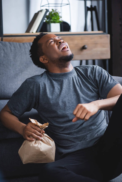 hombre afroamericano estresado sosteniendo bolsa de papel mientras tiene ataque de pánico en casa
 - Foto, Imagen