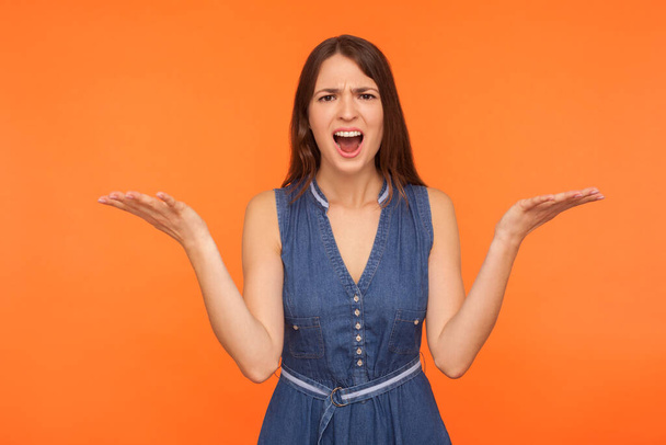 How could you? Annoyed disgruntled brunette girl in denim dress angrily asking reason of failure while quarreling scolding, emotions of irritated indignant woman. indoor studio shot orange background - Foto, afbeelding