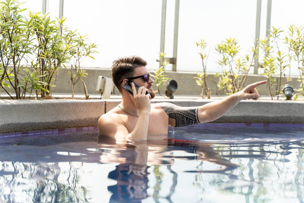 Mladý muž se slunečními brýlemi používající svůj mobil v bazénu, zatímco ukazuje zvednutým prstem - Fotografie, Obrázek