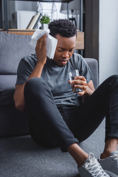 stressed sweaty african american man holding glass of water and napkins at home - Fotó, kép