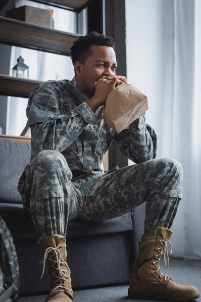 african american soldier in military uniform breathing with paper bag while having panic attack at home - Fotografie, Obrázek