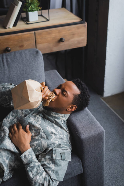 african american soldier in military uniform breathing with paper bag while having panic attack at home - Photo, Image