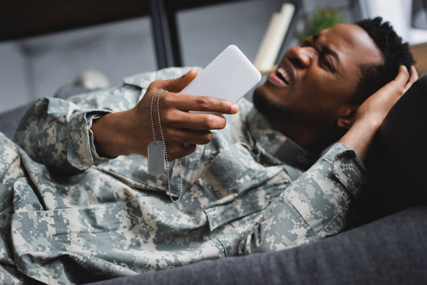 напружений американець - афроамериканець плаче, тримаючи армійський значок і смартфон, страждаючи вдома від ПТСД. - Фото, зображення