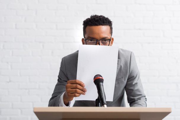 νευρικό αφροαμερικανός ομιλητής κρύβεται πίσω από το χαρτί κατά τη διάρκεια της επιχειρηματικής διάσκεψης - Φωτογραφία, εικόνα