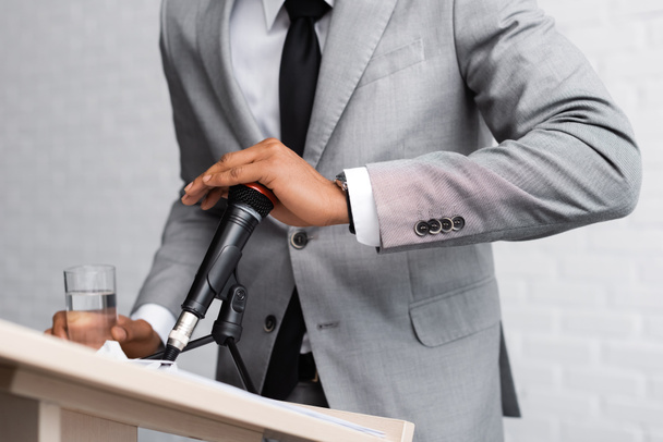 oříznutý pohled na nervózní africký americký reproduktor držící sklenici vody a zakrývající mikrofon během obchodní konference  - Fotografie, Obrázek