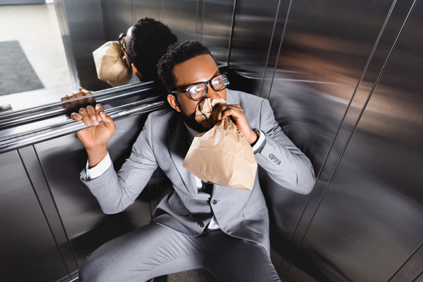 испуганный африканский американский бизнесмен дышит бумажным пакетом и страдает от панической атаки в лифте
 - Фото, изображение