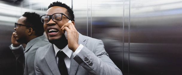 Panoramaaufnahme eines verängstigten weinenden afrikanisch-amerikanischen Geschäftsmannes, der mit dem Smartphone spricht und im Aufzug unter Panikattacken leidet - Foto, Bild