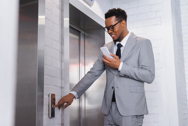 positive african american businessman using smartphone while pushing elevator button - Photo, Image