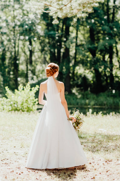 η νύφη με ένα λευκό φόρεμα και ένα πέπλο στα μαλλιά της, στέκεται με την πλάτη μπροστά μας και κρατά ένα μπουκέτο λουλούδια στο φόντο ενός πράσινου δάσους. θολό φόντο - Φωτογραφία, εικόνα