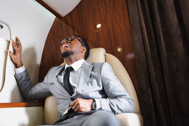 Αφροαμερικάνος αγχωμένος επιχειρηματίας που κρατά ζώνη ασφαλείας και υποφέρει από κρίση πανικού κατά τη διάρκεια της πτήσης με ιδιωτικό αεροπλάνο - Φωτογραφία, εικόνα