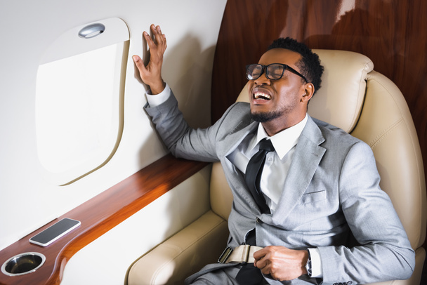Αφροαμερικάνος επιχειρηματίας που κρατά ζώνη ασφαλείας και υποφέρει από κρίση πανικού κατά τη διάρκεια της πτήσης με ιδιωτικό αεροπλάνο - Φωτογραφία, εικόνα