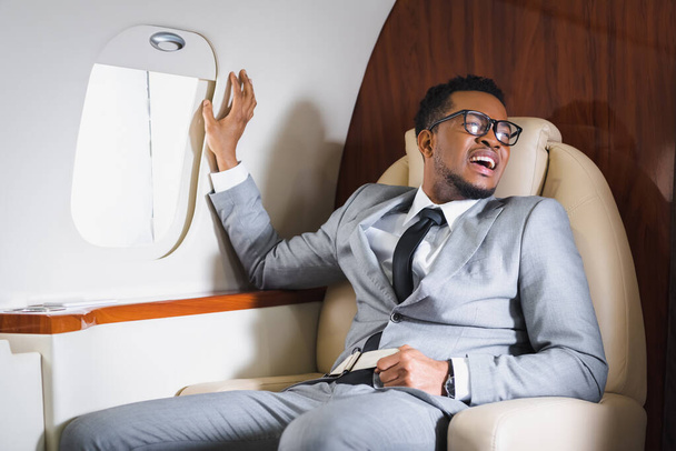 african american businessman holding safety belt and suffering from panic attack during flight on plane - Photo, Image