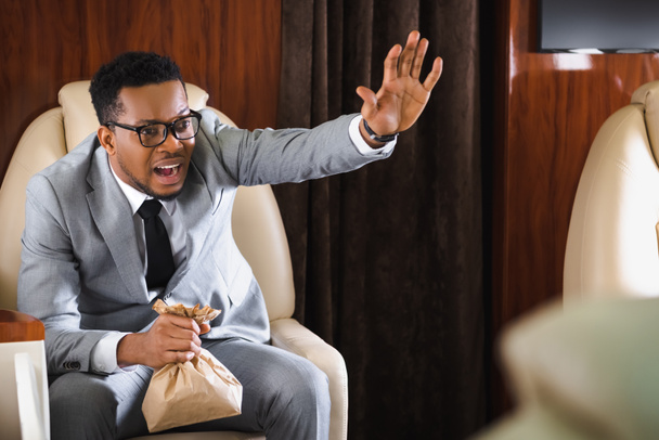 empresario afroamericano estresado con bolsa de papel teniendo ataque de pánico durante el vuelo y mostrando gesto de parada
 - Foto, Imagen
