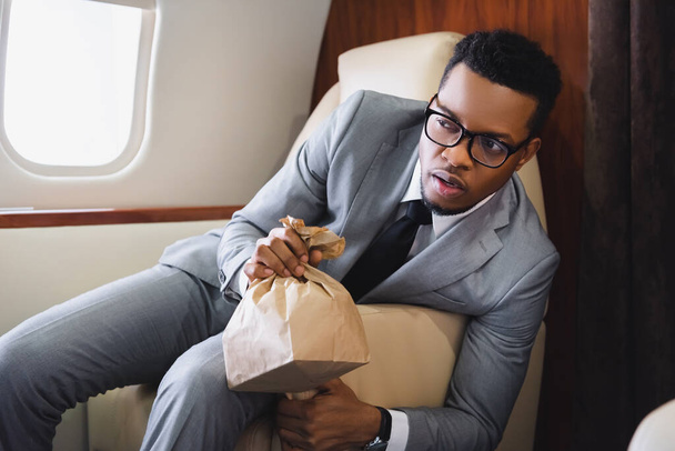 Homme d'affaires afro-américain inquiet tenant un sac en papier tout en ayant une attaque de panique pendant le vol en avion privé
 - Photo, image