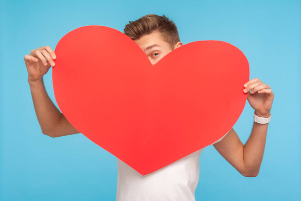 Romantic congratulation on Valentine day! Young man peeking out of big red paper heart, flirting, looking playfully to camera, declaration of love. indoor studio shot isolated on blue background - Photo, Image