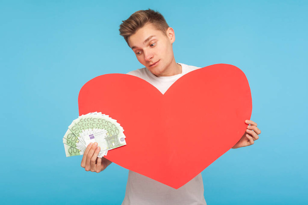 Portrait of unhappy young rich man holding big red heart and looking sadly at euros in his hand, disappointed in mercenary love, dating for money. indoor studio shot isolated on blue background - Zdjęcie, obraz