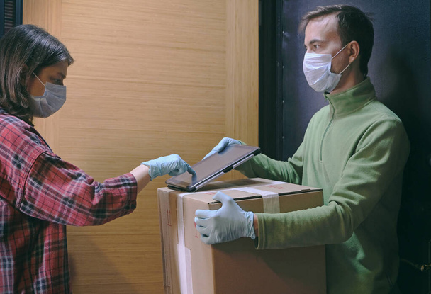 Courier in protective mask delivers parcel, customer in medical gloves signs on tablet. Delivery service under quarantine, disease outbreak, coronavirus covid-19 pandemic conditions. - Photo, image