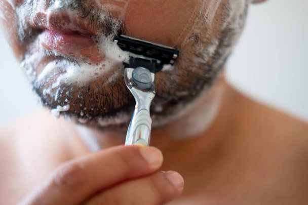 Joven con jabón de espuma en las mejillas está de pie y quitándose el bigote afeitándose la cara
. - Foto, imagen
