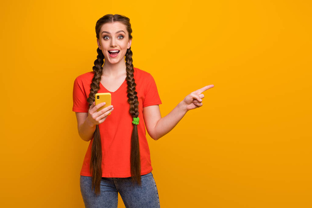 Photo of pretty funny teen lady hold telephone millennial blogger direct finger side empty space open mouth wear casual red t-shirt jeans isolated bright yellow color background - Photo, Image