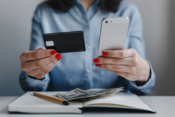 Kırmızı tırnaklı, elinde kredi kartı ve telefon olan mavi gömlekli bir kadının masanda oturup ödemesini yaptığı fotoğraf. İş, serbest meslek, uzak iş, online iş kavramı - Fotoğraf, Görsel
