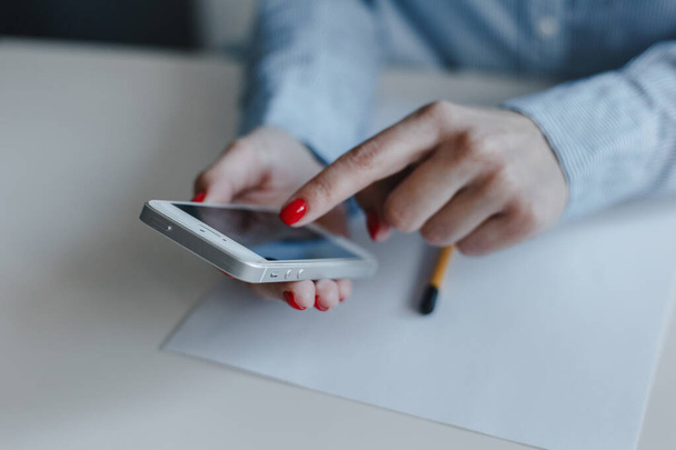 Zbliżenie rąk kobiety z czerwonymi paznokciami trzymając i klikając telefon komórkowy dokonywania płatności online i czatując w niebieskiej koszuli. Biznes, wolny strzelec, praca na odległość, koncepcja pracy online - Zdjęcie, obraz