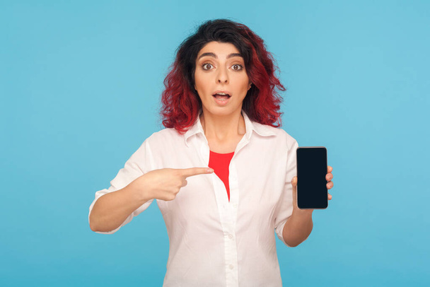 Portrait of amazed shocked hipster woman with fancy red hair pointing to smartphone and looking with surprised big eyes, recommending excellent mobile service. studio shot isolated on blue background - Foto, Imagem