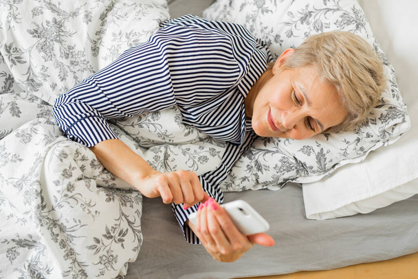 красива літня жінка в піжамі на ліжку під обкладинками з телефоном
 - Фото, зображення