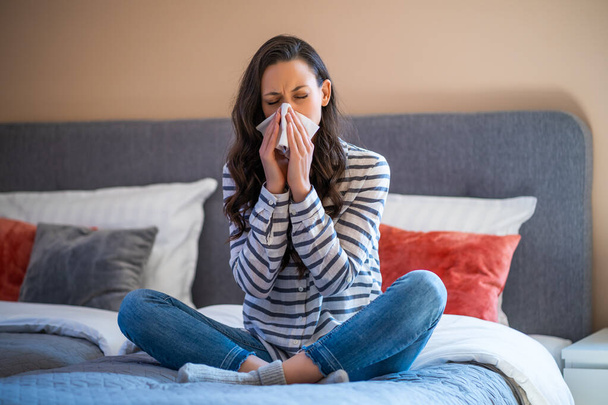 Γυναίκα κάθεται στο κρεβάτι στο σπίτι της, έχει γρίπη και φυσάει μύτη. - Φωτογραφία, εικόνα