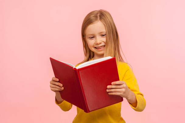 Smart positive cute little girl reading big red book and smiling happily, learning homework, being curious about school knowledge, education concept. indoor studio shot isolated on pink background - Photo, Image