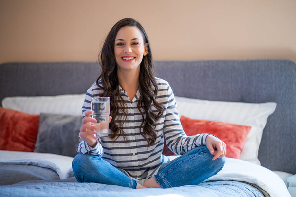Šťastná mladá žena sedí na posteli ve svém domě, dívá se na kameru a drží sklenici vody. - Fotografie, Obrázek