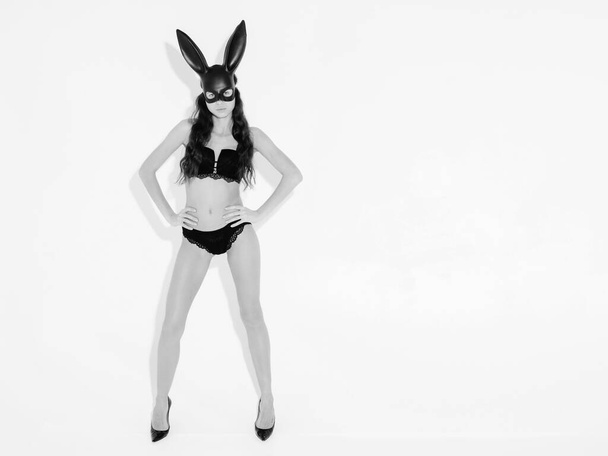 Beautiful sexy woman with large breasts wearing  carnival black mask of Easter bunny rabbit.Hot blonde girl posing near white wall in studio. Seductive model in nice lingerie. Black and white - Valokuva, kuva