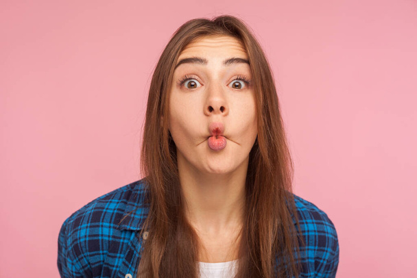 Closeup of amazed childish cute girl in checkered shirt standing with pout lips making fish face, having fun, showing surprised silly expression. indoor studio shot isolated on pink background - 写真・画像