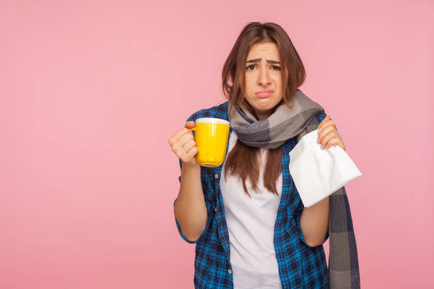 Treatment of seasonal influenza. Portrait of sick girl wrapped in scarf looking with funny displeased grimace, holding tea cup treating flu, cough and fever. studio shot isolated on pink background - Zdjęcie, obraz