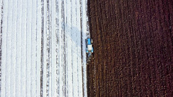 Синий трактор вспахивает поле, покрытое снегом. За трактором чернозем. Россия, Урал, Взгляд с высоты птичьего полета   - Фото, изображение