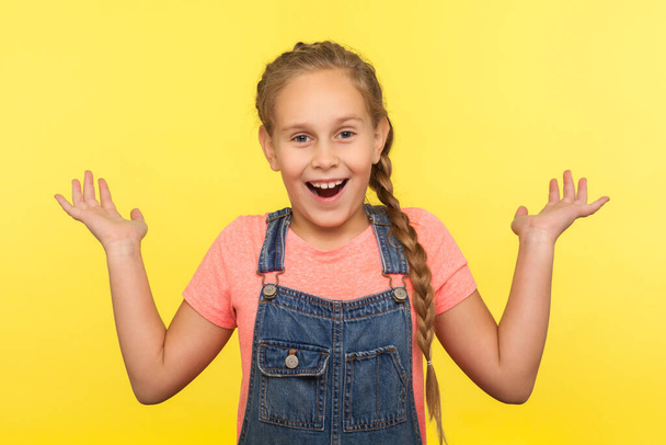 Unexpected surprise. Portrait of astonished child in denim overalls standing with raised hands in amazement, shocked by sudden unbelievable news. indoor studio shot isolated on yellow background - Foto, afbeelding