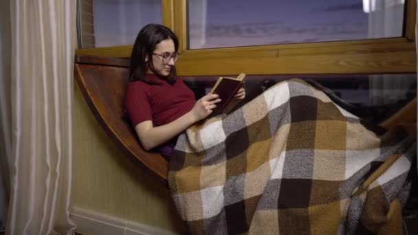 Een jonge vrouw leest een boek. Een meisje ligt op een vensterbank bij het raam met een boek in haar handen. De avond uit. - Video