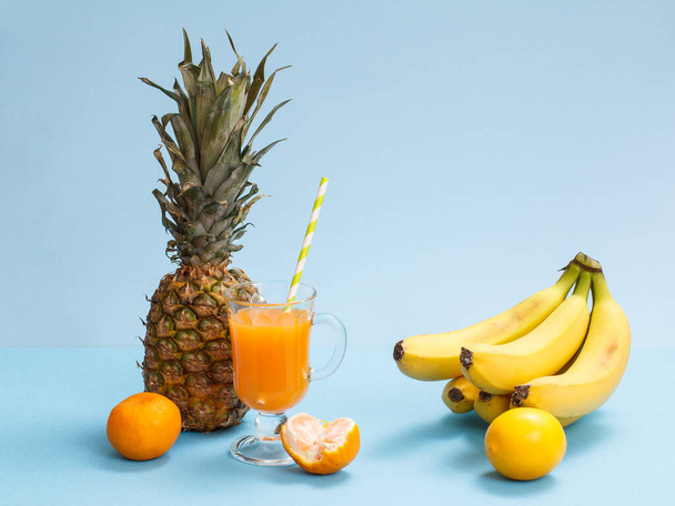 Natural composition from tropical fruits. Fresh pineapple, bananas, tangerines and lemon with glass of fruit juice on blue background. - Photo, Image