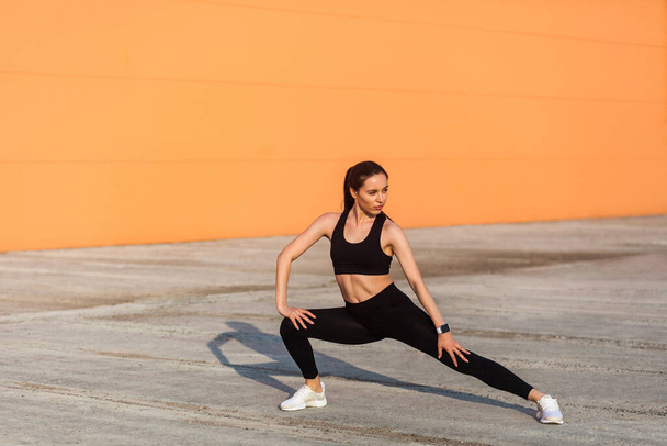 Slim young pretty woman in tight sportswear, black pants and top, practicing outdoor, doing stretching workouts for better flexibility, lower-body exercise. Health care concept, sport activity - Foto, Imagem