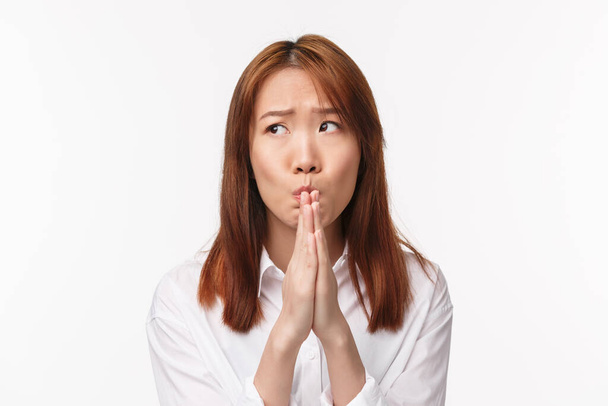 Nahaufnahme Porträt einer ängstlichen nervösen jungen asiatischen Frau in weißem Hemd, die besorgt dasteht, die Hände im Gebet hält, ängstlich wegschaut, die Stirn runzelt, Wünsche wahr werden lässt, flehentlich, weißer Hintergrund - Foto, Bild