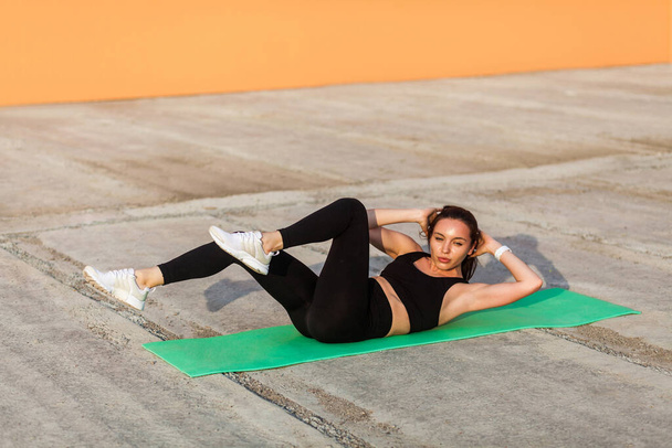 Fit sporty beautiful girl in tight sportswear, black pants and top, lying on mat doing sit ups, crunch exercise, training abdominal muscles. Health care and weight loss concept, sport activity outdoor - Foto, Bild