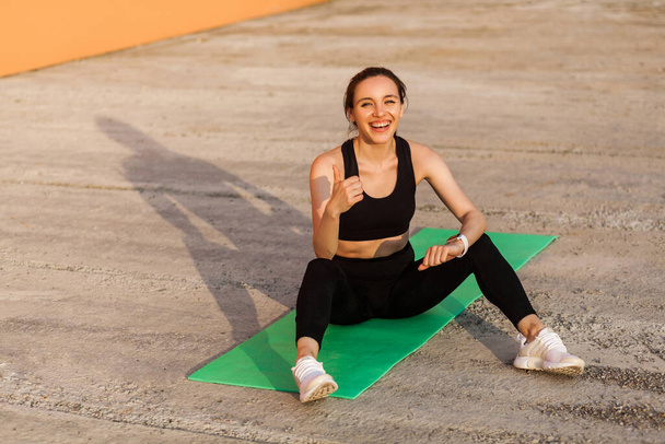 Delighted athletic girl in black pants and top, smiling and showing thumbs up, sitting on yoga mat after workouts, gesturing like, satisfied with physical training. Health care, sport activity outdoor - Zdjęcie, obraz