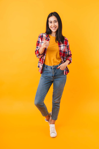 Photo of joyful young woman in casual wear showing thumb up and smiling isolated over yellow background - Fotó, kép