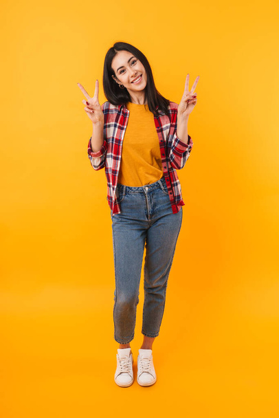 Photo of joyful young woman gesturing peace sign and smiling isolated over yellow background - Photo, image