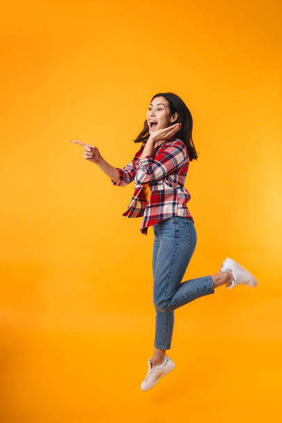 Photo of amazed young woman jumping and pointing finger aside isolated over yellow background - Фото, изображение