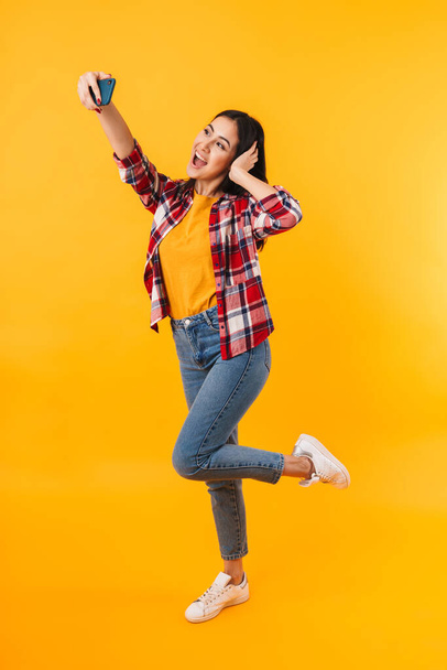 Photo of amazed young woman smiling while taking selfie on cellphone isolated over yellow background - 写真・画像