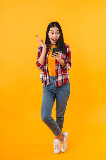 Photo of excited brunette woman expressing surprise while using cellphone isolated over yellow background - Photo, Image
