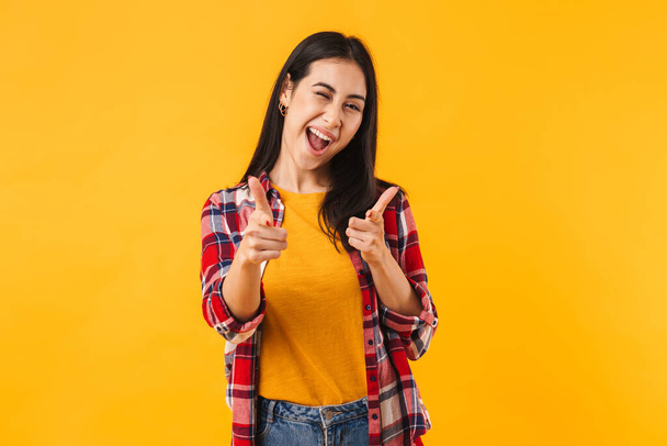 Photo of joyful brunette woman winking while pointing fingers at camera isolated over yellow background - Photo, image