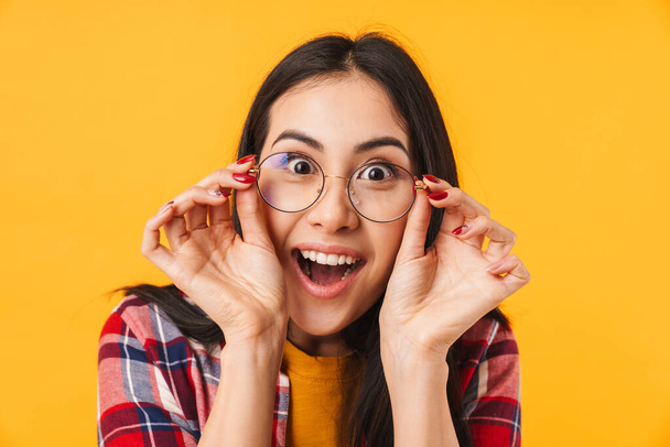 Photo of surprised brunette woman holding eyeglasses and looking at camera isolated over yellow background - Fotografie, Obrázek